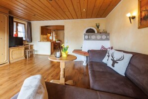 A cozy living room with a wooden ceiling, a brown sofa with a deer pillow, a round wooden table, and a television on a wooden cabinet. - Holiday apartment with 2 bedrooms (Ruhpolding)
