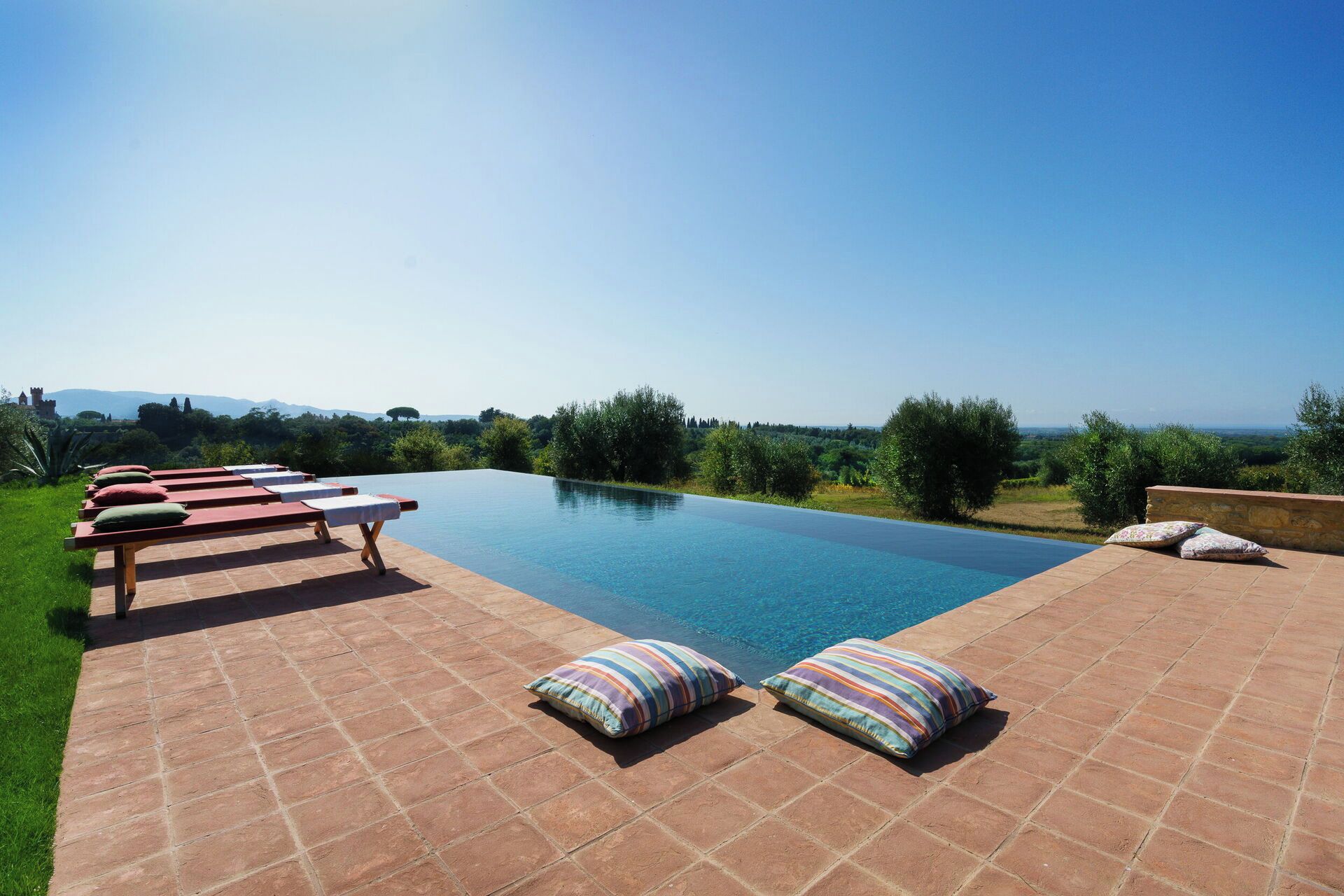 An infinity pool with lounge chairs and pillows, surrounded by greenery and a clear sky.