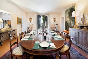 A formal dining room with a long table set for a meal, surrounded by chairs, and a view of the outdoors through a doorway. - Villa Bellaria, Private Pool, Bolgheri (Bolgheri)