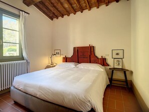 A bedroom with a wooden headboard, a bed with white linens, a nightstand with a lamp, a radiator, and a window with curtains. - Villa Bellaria, Private Pool, Bolgheri (Bolgheri)