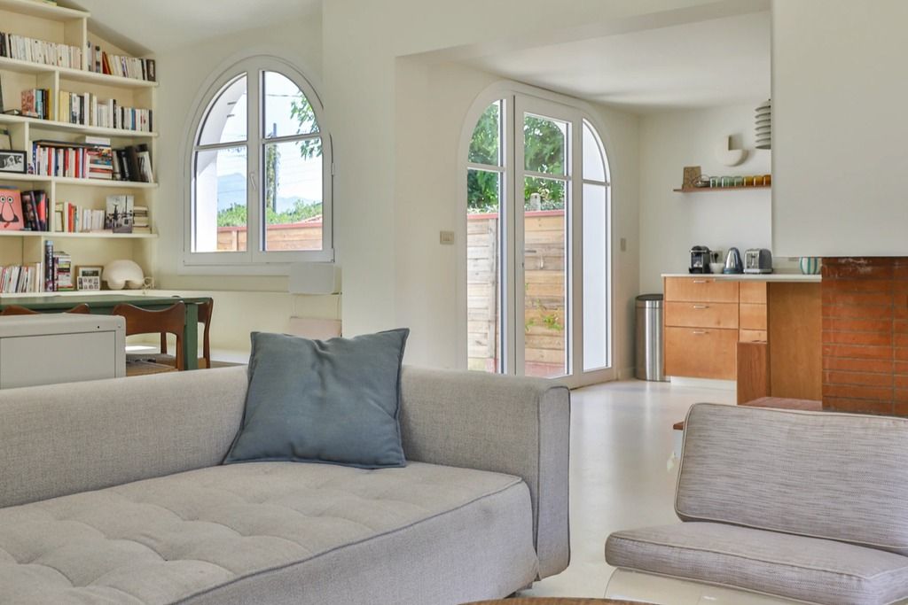 A modern living room with a sofa, a bookshelf, and a kitchen area visible through an arched window.