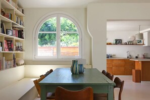 Ein Essbereich mit großem Fenster, einer Bücherwand und einer Küche im Hintergrund.
