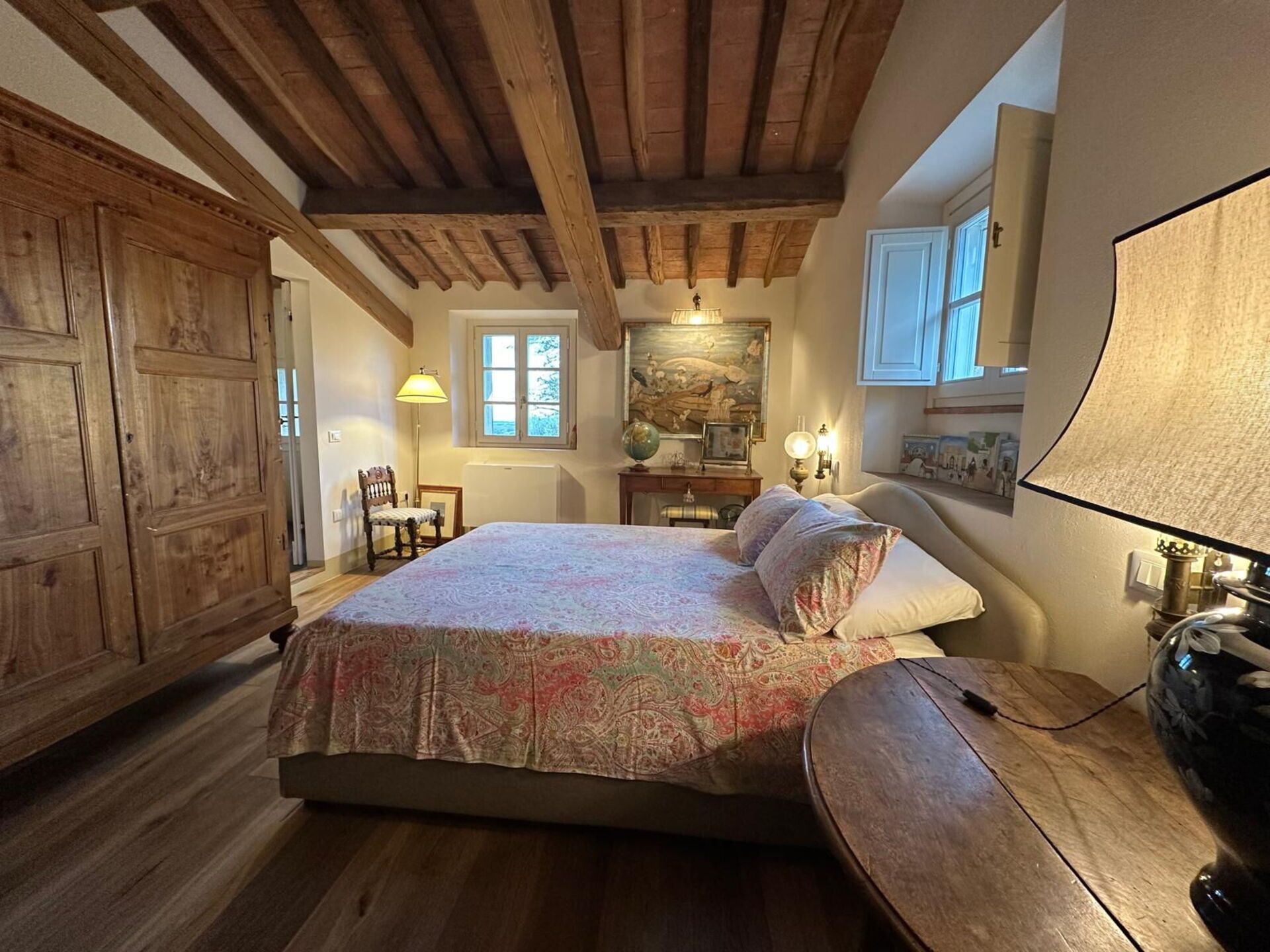 A bedroom with a wooden ceiling, a bed with patterned bedding, a desk with a lamp, and a large window.