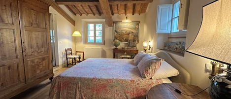 A bedroom with a wooden ceiling, a bed with patterned bedding, a desk with a lamp, and a large window.