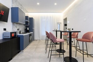 A modern kitchen with dark cabinetry, a built-in oven, and a coffee machine. There is a dining area with a round table and pink chairs. - Vittorio Emanuele Arte Suites Guesthouse (Rome)