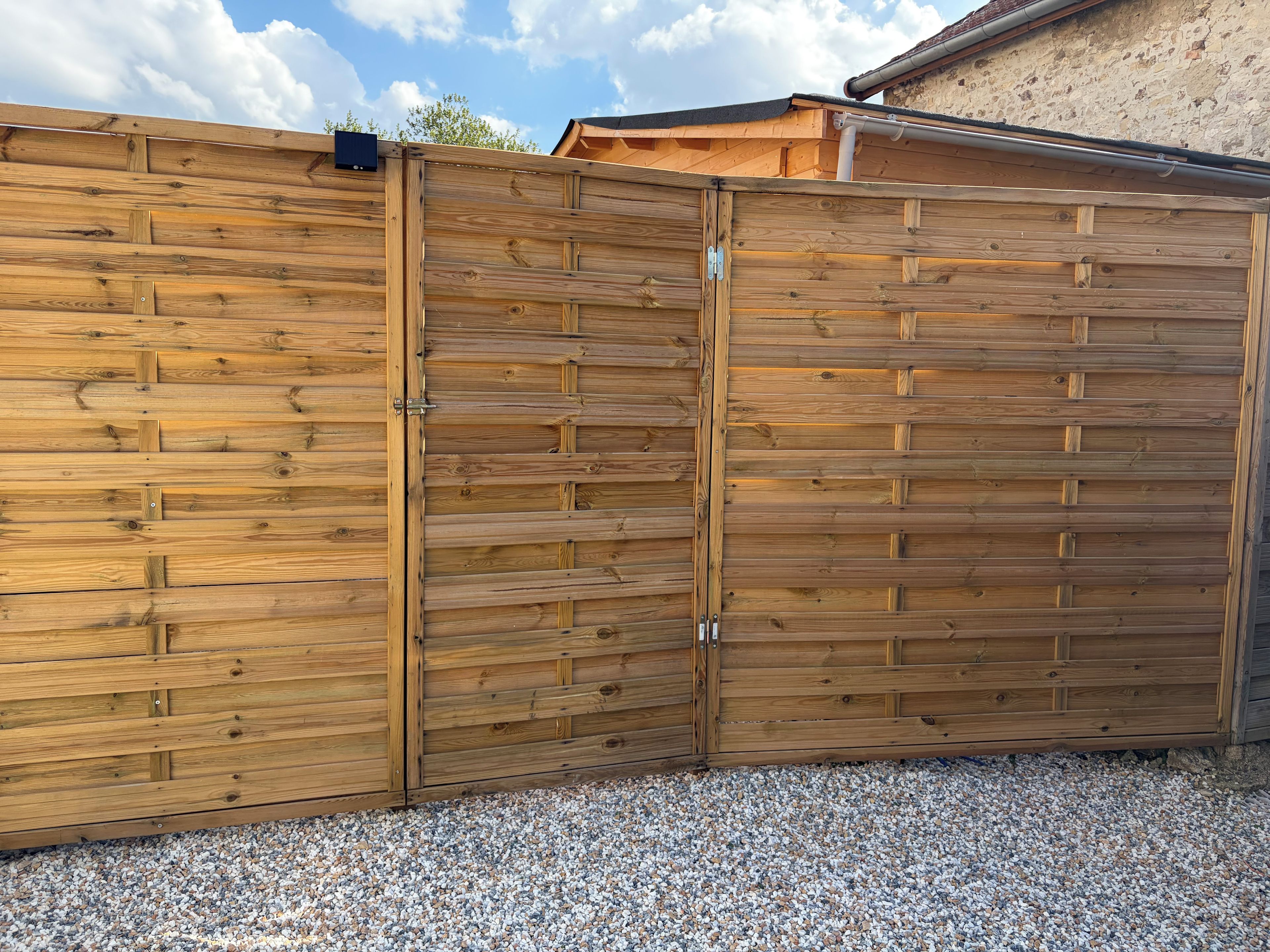 A wooden fence with a gate, gravel ground, and a building in the background.