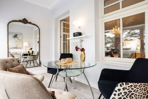Ein modernes Wohnzimmer mit einem Glastisch, einer Couch und einem Sessel. An der Wand befindet sich ein großer Spiegel, ein Heizkörper und ein Fenster mit Blick ins Freie.