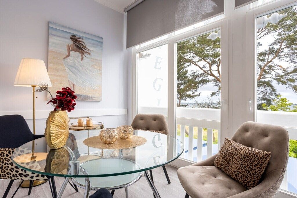 A modern living room with a glass table, comfortable seating, a painting on the wall, and a view through large windows.