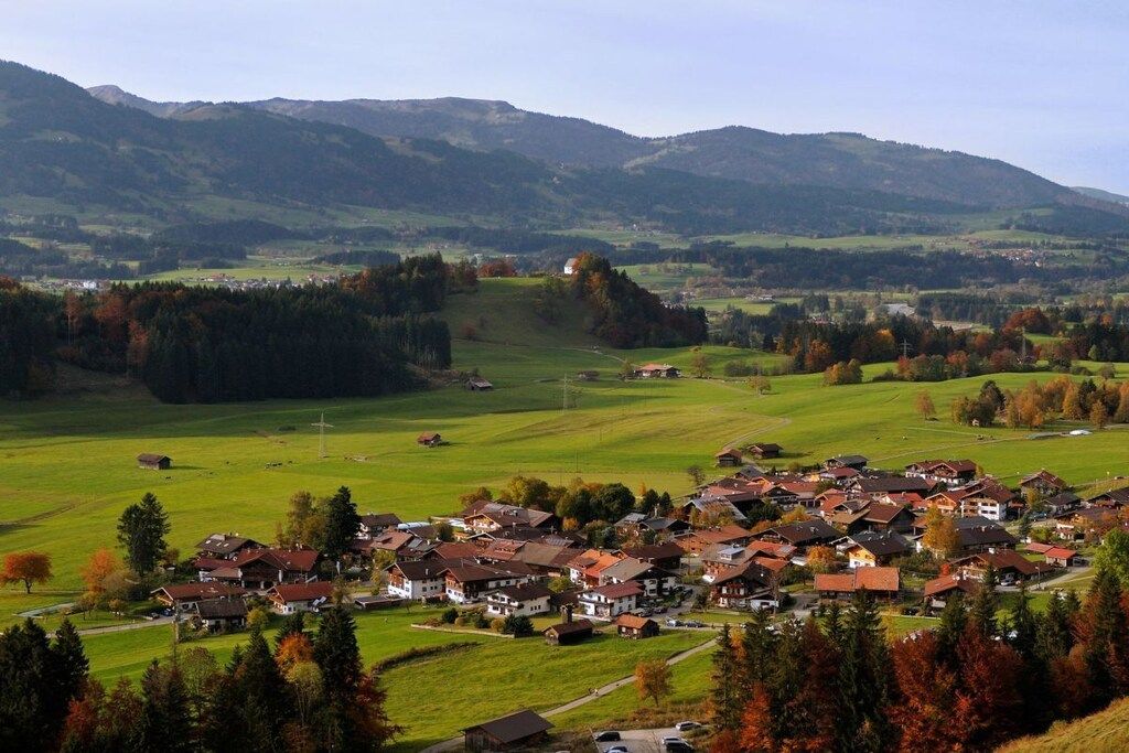 Ein Dorf mit Häusern und einer Kirche, umgeben von grünen Feldern und Bergen.