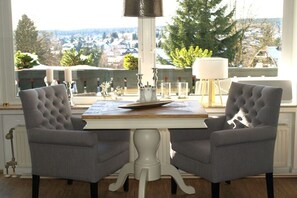 A dining area with a table and two chairs, a lamp, and a view of a town through large windows. - Holiday apartment with 1 bedroom (Braunlage)