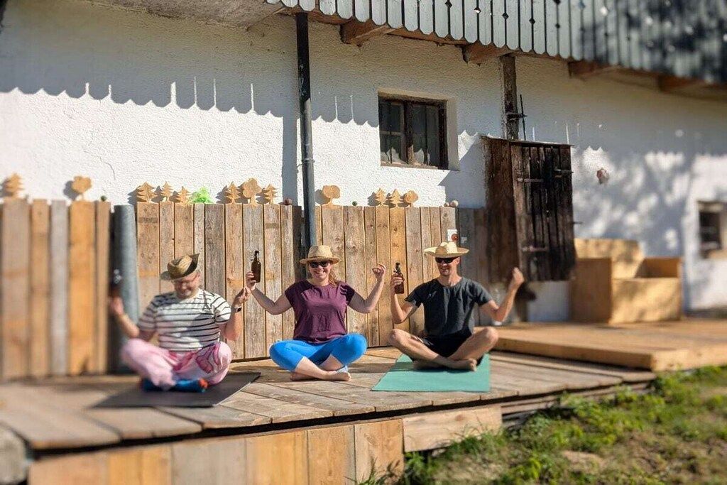 Three individuals seated on mats outdoors with a wooden deck and a white building with a window in the background.