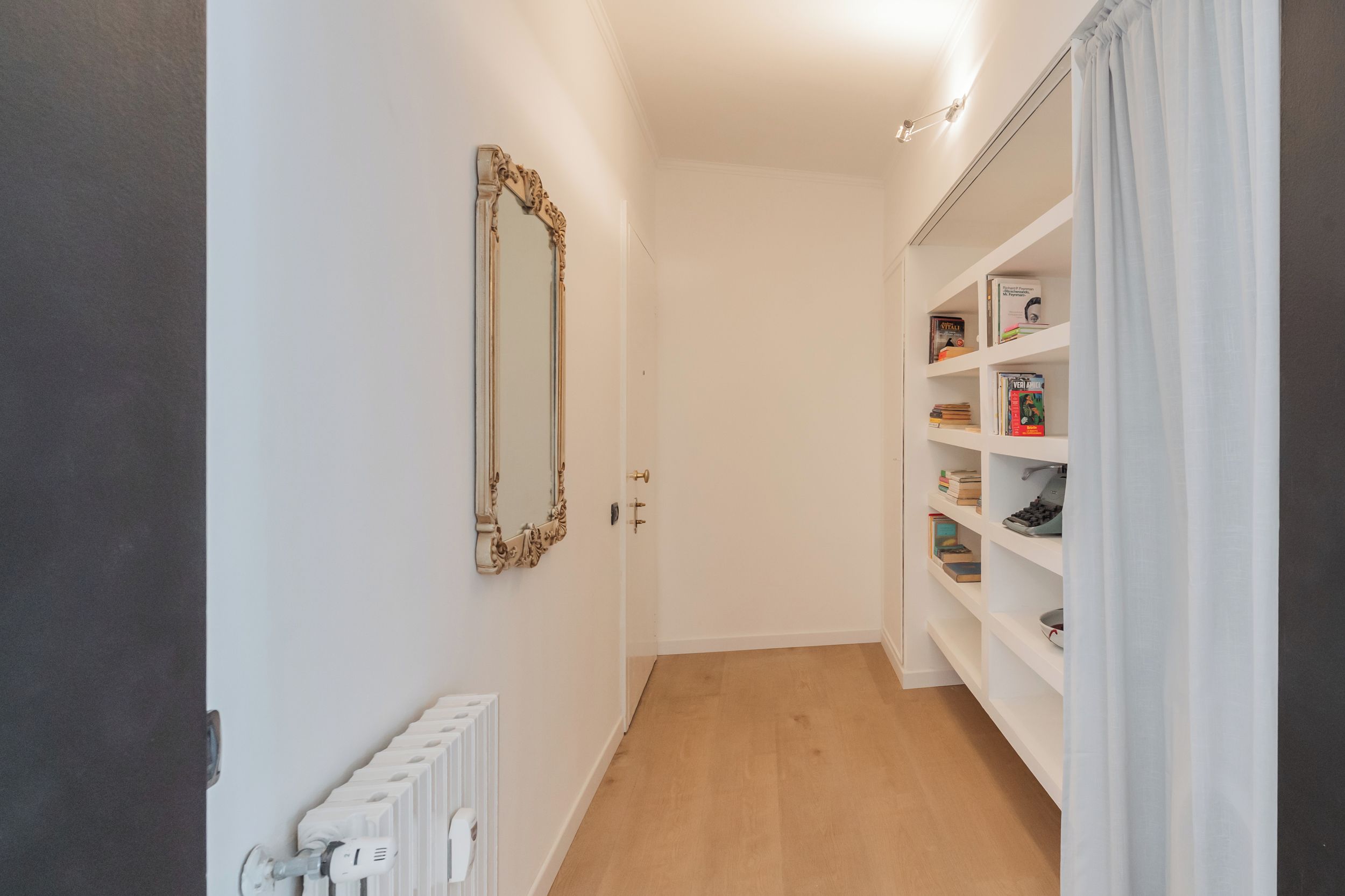 A narrow hallway with a radiator, a mirror, and a bookshelf.