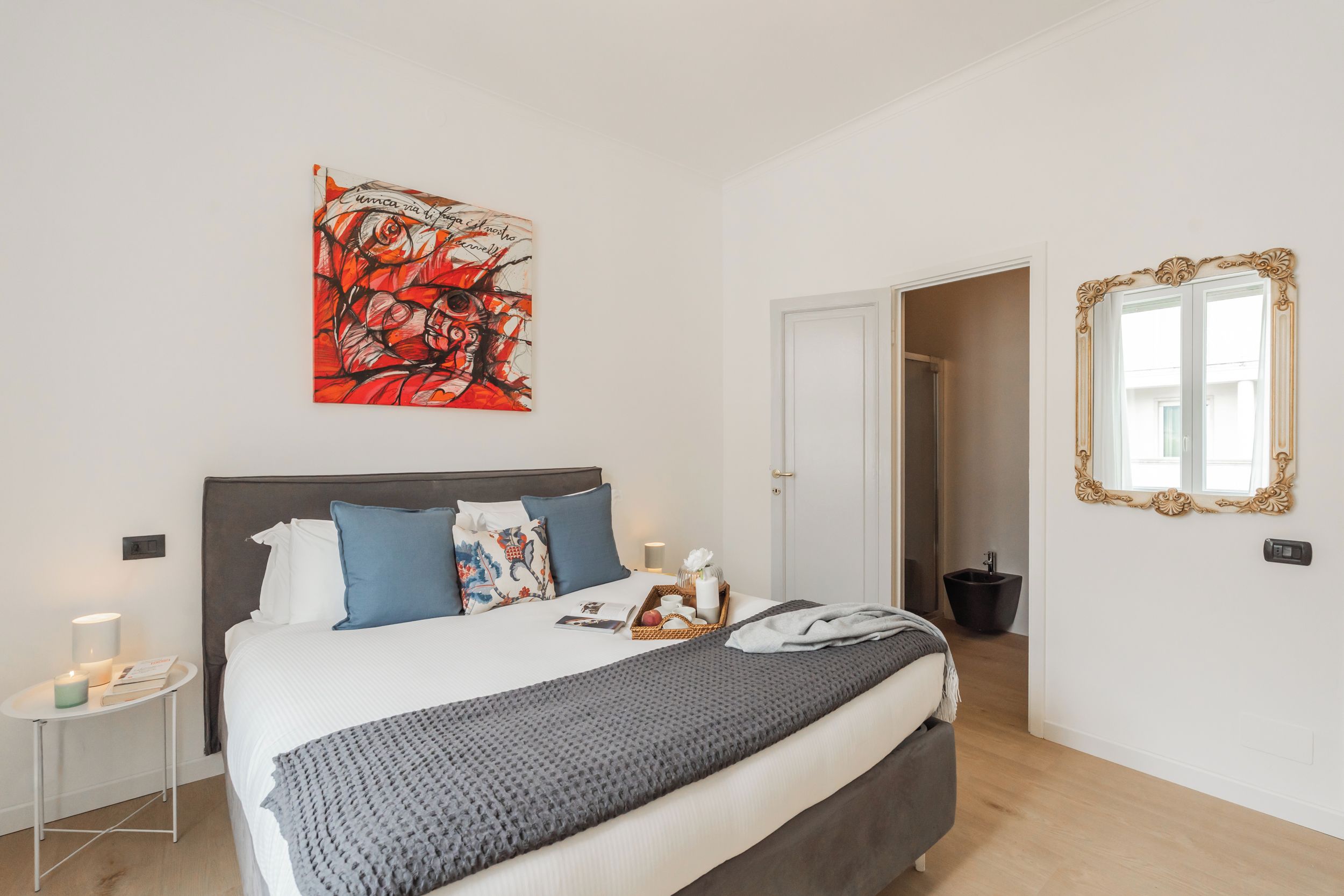 A bedroom with a bed, pillows, a bedside table with a lamp, a wall art piece, a mirror, and a door leading to another room.
