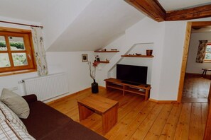 A living room with a wooden floor, a brown sofa, a TV on a wooden stand, and a window with curtains. - Holiday home with 1 bedroom (Neuschönau)
