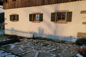 A building with a white exterior, wooden shutters, and a stone pathway with snow. - Holiday home with 1 bedroom (Neuschönau)