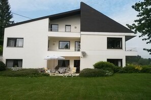 Ein modernes, zweistöckiges Gebäude mit flachem Dach, einem Balkon und einer Terrasse mit Gartenmöbeln.