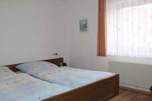 Un dormitorio con una cama de madera, una pared blanca, un ventanal con cortinas traslúcidas y un pequeño cuadro enmarco.