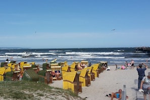 Ein Strand mit Reihen von Liegestühlen und Sonnenschirmen, teilweise besetzt mit Personen, und einem Steg im Hintergrund.
