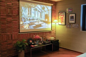 A cozy room with a brick wall, a projection screen displaying a room, a small table with a lamp, and a few framed items on the wall. - Benri Homestay (Cau Kieu)