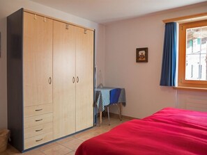 A bedroom with a large wardrobe, a bed with a red cover, a small table with a checkered tablecloth, and a window with blue curtains. - Heidblick Appartement/Fewo, Dusche oder Bad, WC 3½-Zimmerwohnung by Interhome (Klosters)