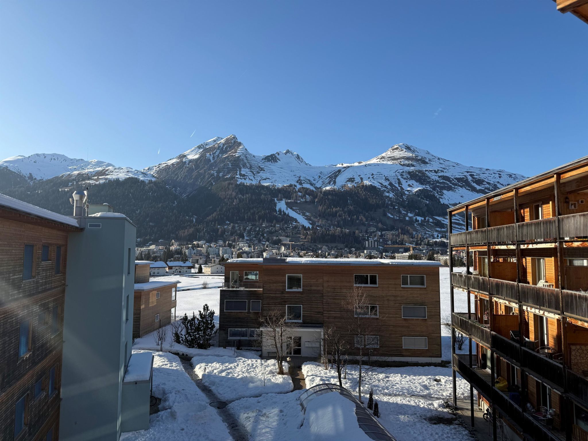 Ein modernes Mehrstöckgebäude, verschneiter Boden und ein Gebirge im Hintergrund.