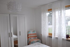 Ein Schlafzimmer mit einem Bett, einem Stuhl mit gemustertem Bezug, einem Fenster mit transparenten Vorhängen und einem Kleiderschrank.