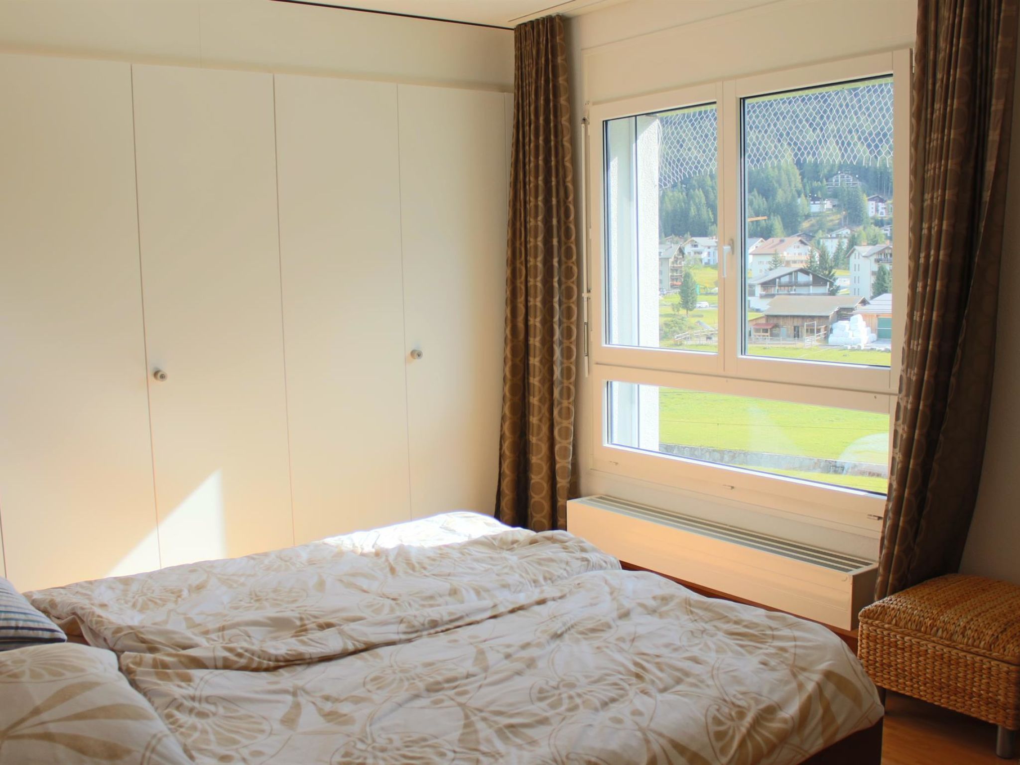 A bedroom with a bed, a window with curtains, and a view of a residential area.