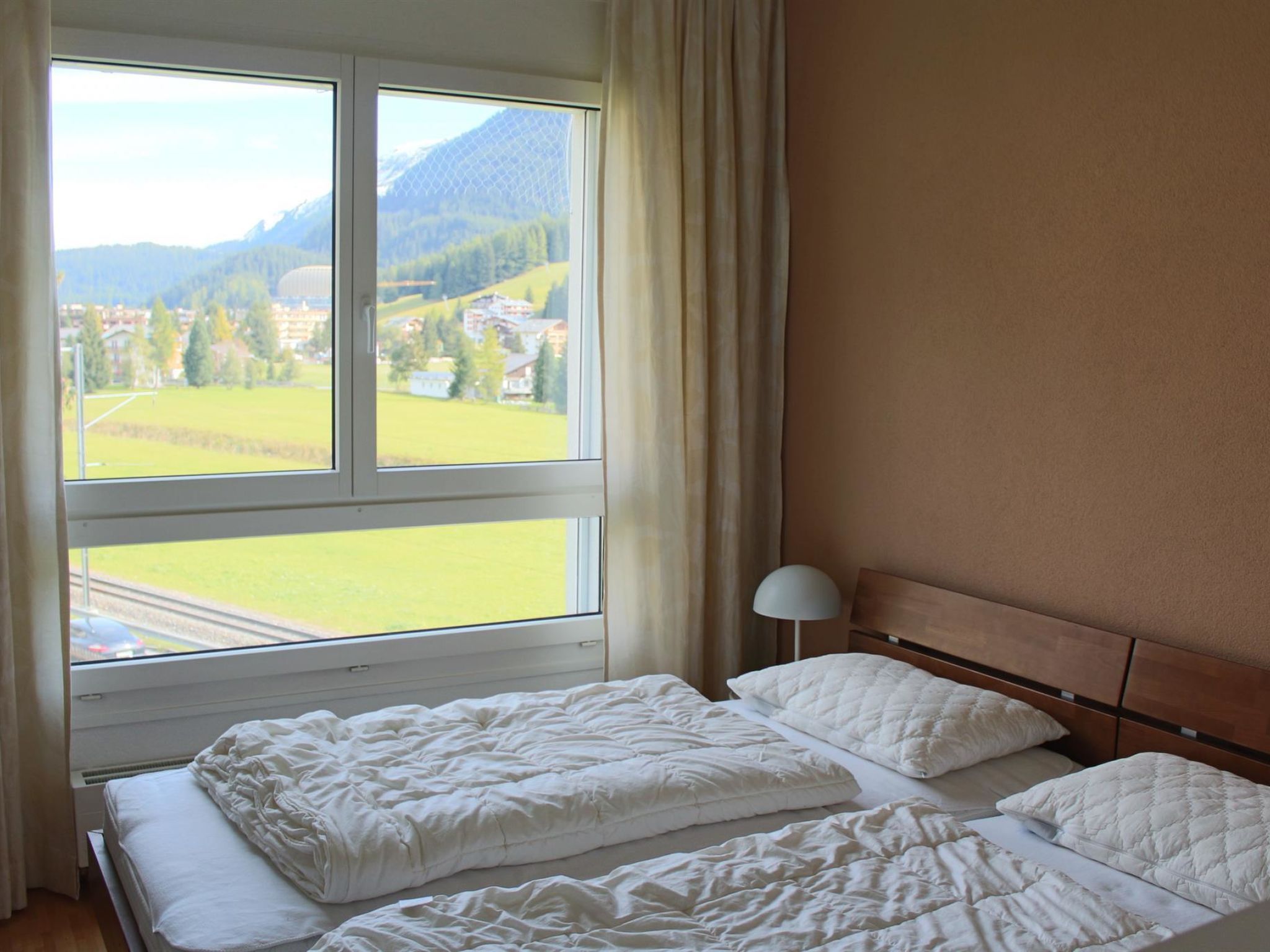 Une chambre d’hôtel avec deux lits, une fenêtre donnant sur un paysage verdoyant et des bâtiments, et une lampe sur la table de chevet.