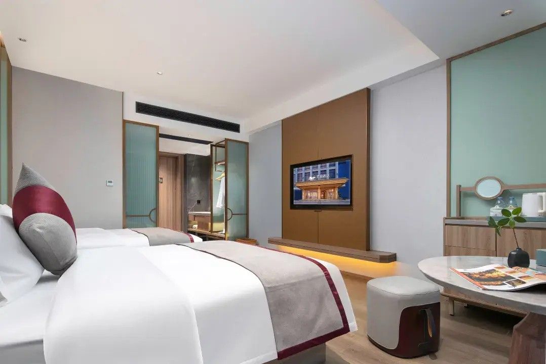 A modern hotel room with two beds, a sitting area, a television, and a bathroom visible through an open door.
