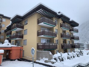 Een meerlaags gebouw met balkons, een met sneeuw bedekte grond en een bergachtige achtergrond.