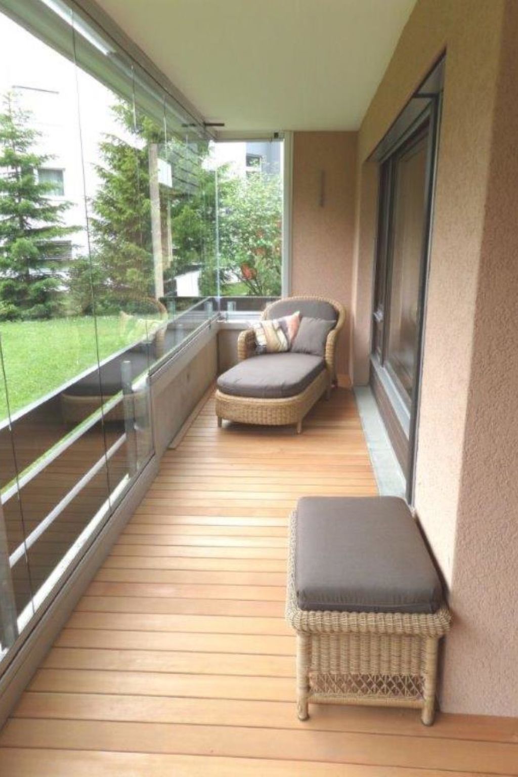 A balcony with a wicker chair and ottoman, glass railings, and a view of greenery.