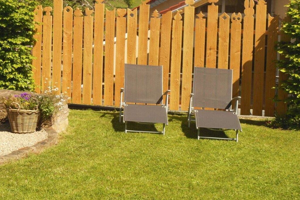 Deux fauteuils de jardin, avec une structure en métal et un revêtement en tissu gris, sont placés sur une pelouse bien entretenue, avec une clôture en bois en arrière-plan.