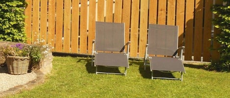 Two outdoor lounge chairs with metal frames and grey fabric seating, positioned on a well-maintained lawn with a wooden fence in the background.