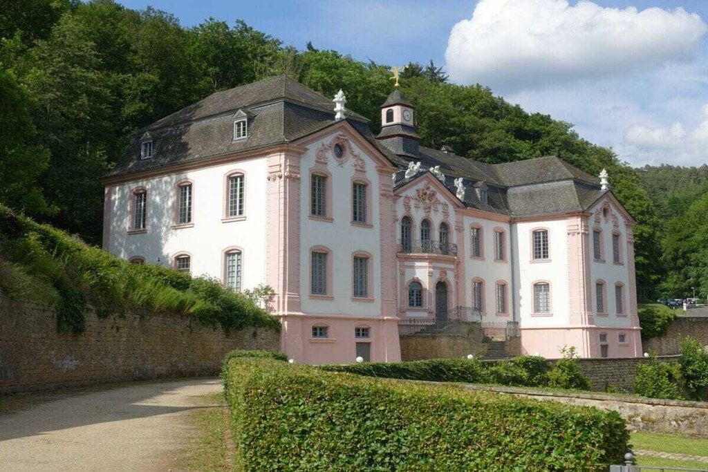 Un grand bâtiment rose et blanc, doté de nombreuses fenêtres et surmonté d’une tour, le tout entouré de verdure.