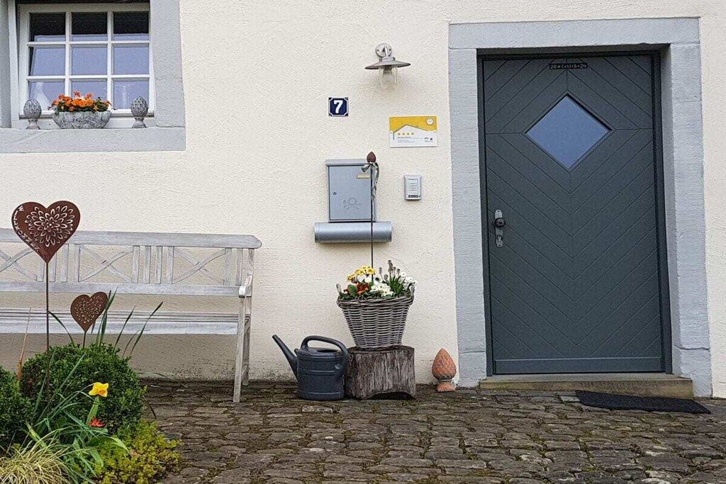 A building facade with a numbered door, a bench, a potted plant, and a watering can.