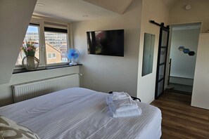 Un dormitorio con cama, televisor de pantalla plana, ventana con vistas a edificios y ventilador.
