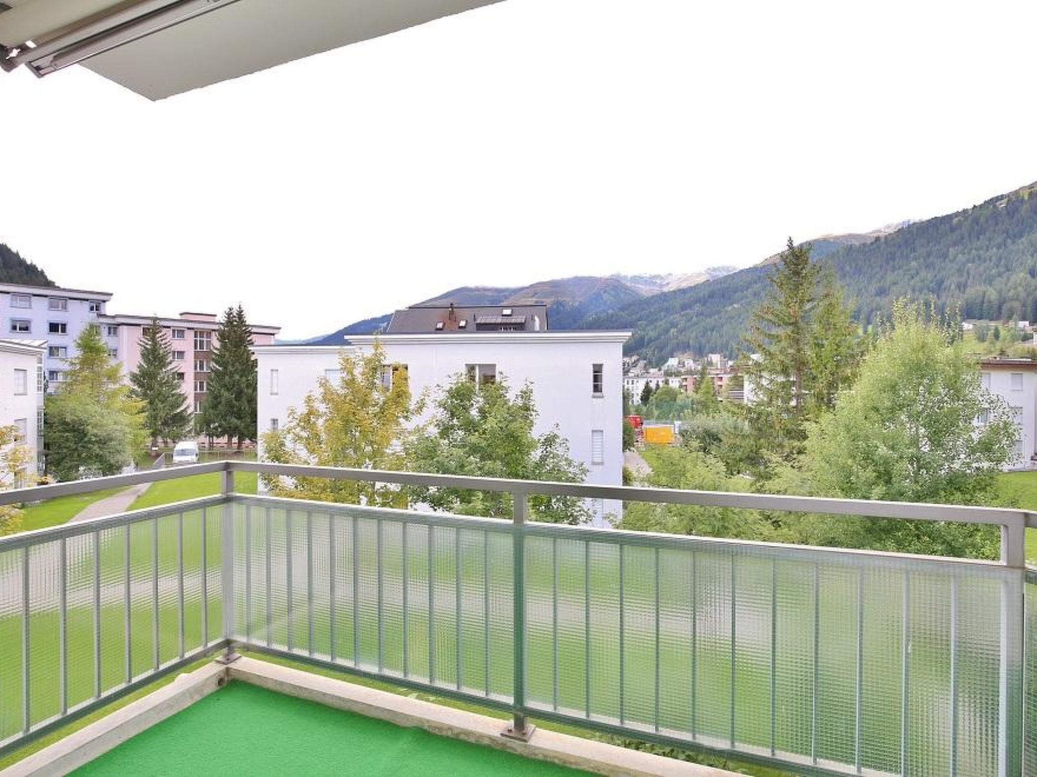 Uma varanda com piso de grama sintética verde, com vista para uma área residencial com edifícios e uma montanha ao fundo.