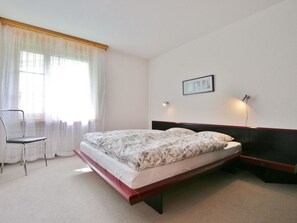 A bedroom with a bed, a chair, a window with curtains, and a wall-mounted light fixture. - Auf der Matta C22 Appartement/Fewo, Dusche oder Bad, WC 3-Zimmerwohnung by Interhome (Davos)