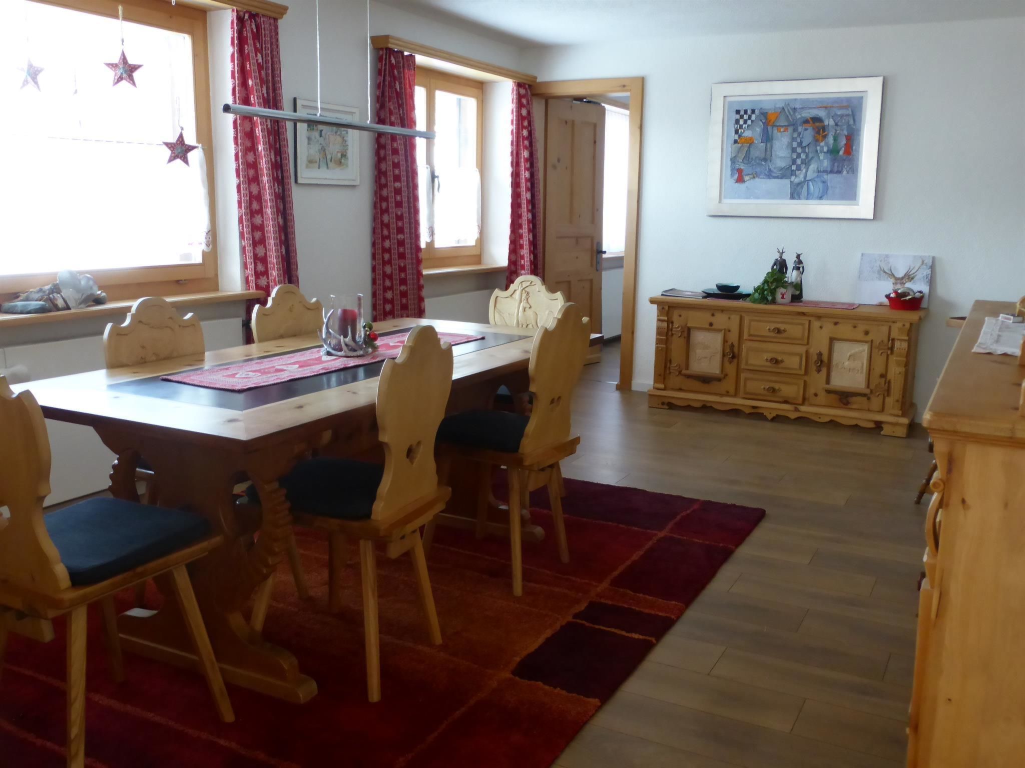 Zona de dinar amb taula de fusta, cadires i una catifa vermella. Hi ha un armari amb objectes decoratius i un quadre a la paret.