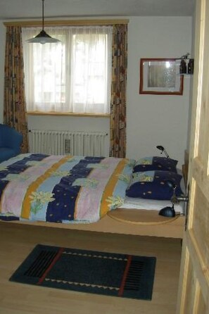 Ein Schlafzimmer mit einem Bett, einem Heizkörper, einem Fenster mit Vorhängen, einem Schreibtisch mit Stuhl und einem Bilderrahmen an der Wand.