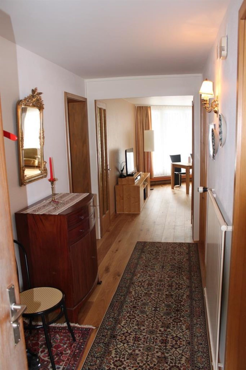 A hallway with a wooden floor, a patterned rug, a mirror, a dresser, and a chair.