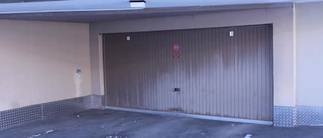 Un espace de stationnement clos par une barrière métallique, avec une porte en métal, un panneau indiquant l’interdiction de se garer et un panneau portant le chiffre « 1 » sur le mur.