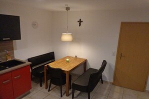 Eine kleine Küche mit Spüle, einem Holztisch und einer Bank. An der Wand hängt eine Wanduhr und ein Kruzifix.