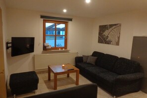 Ein Wohnzimmer mit einer Couch, einem Couchtisch, einem an der Wand befestigten Fernseher und einem Fenster mit Blick auf ein Gebäude.