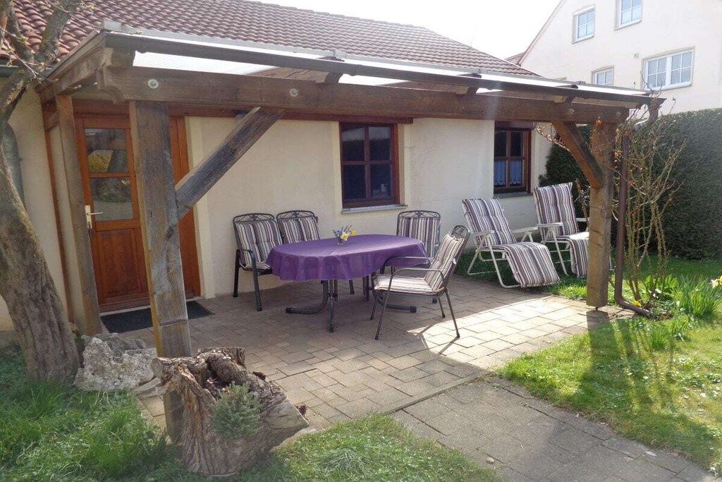 Eine überdachte Terrasse mit Tisch und Stühlen, eine Holz-Pergola und ein Haus im Hintergrund.