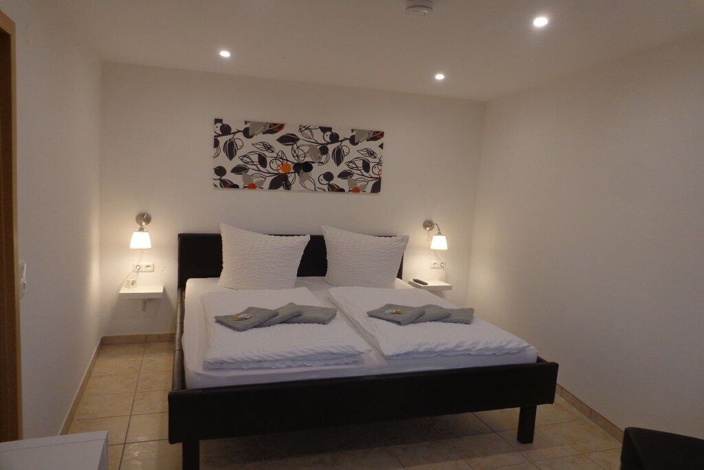 A neatly made double bed with white linens and pillows, two bedside lamps, a wall-mounted artwork, and a tiled floor.