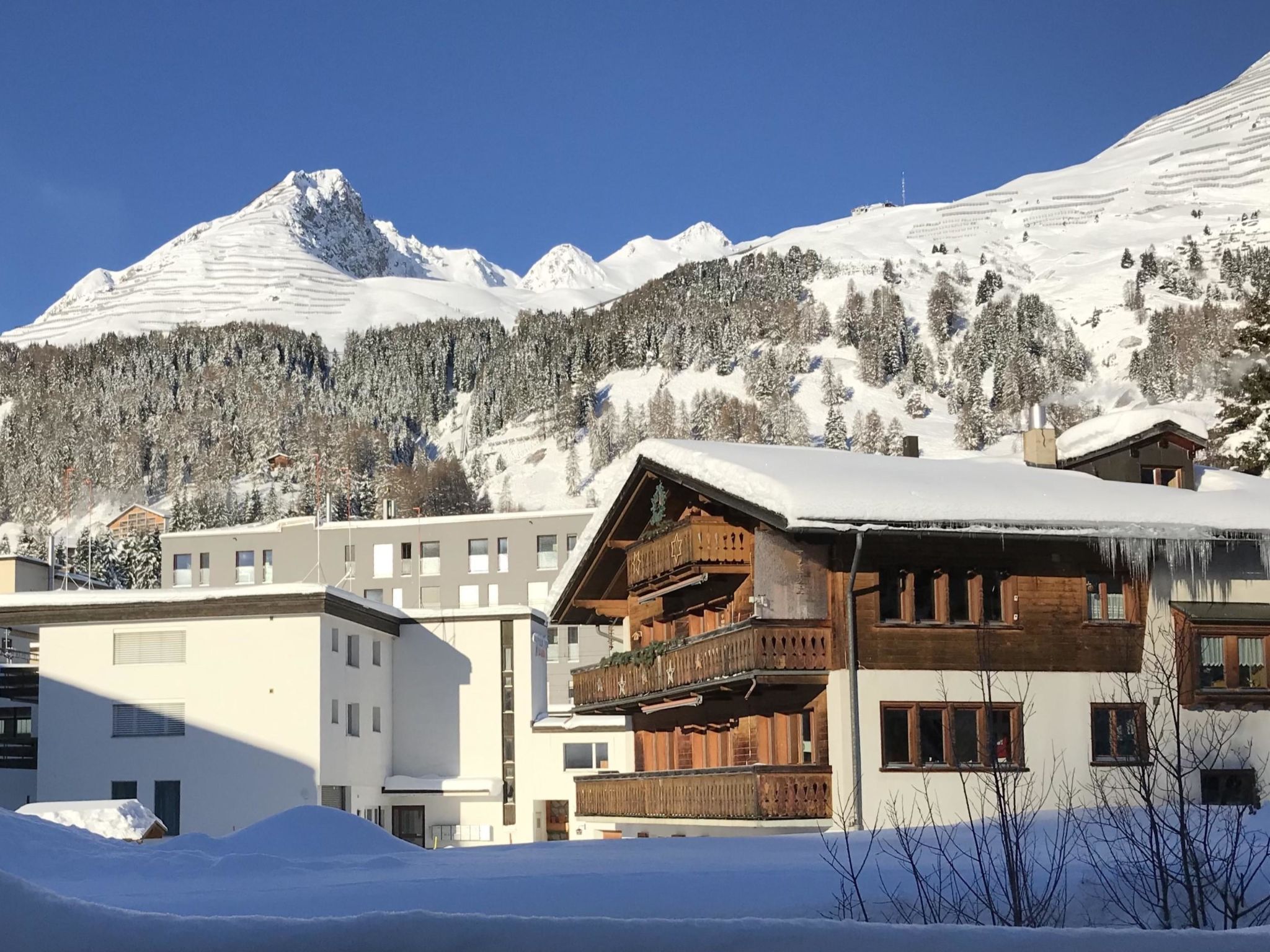 Une montagne enneigée, avec un bâtiment en bois doté de balcons, et un bâtiment moderne en premier plan.