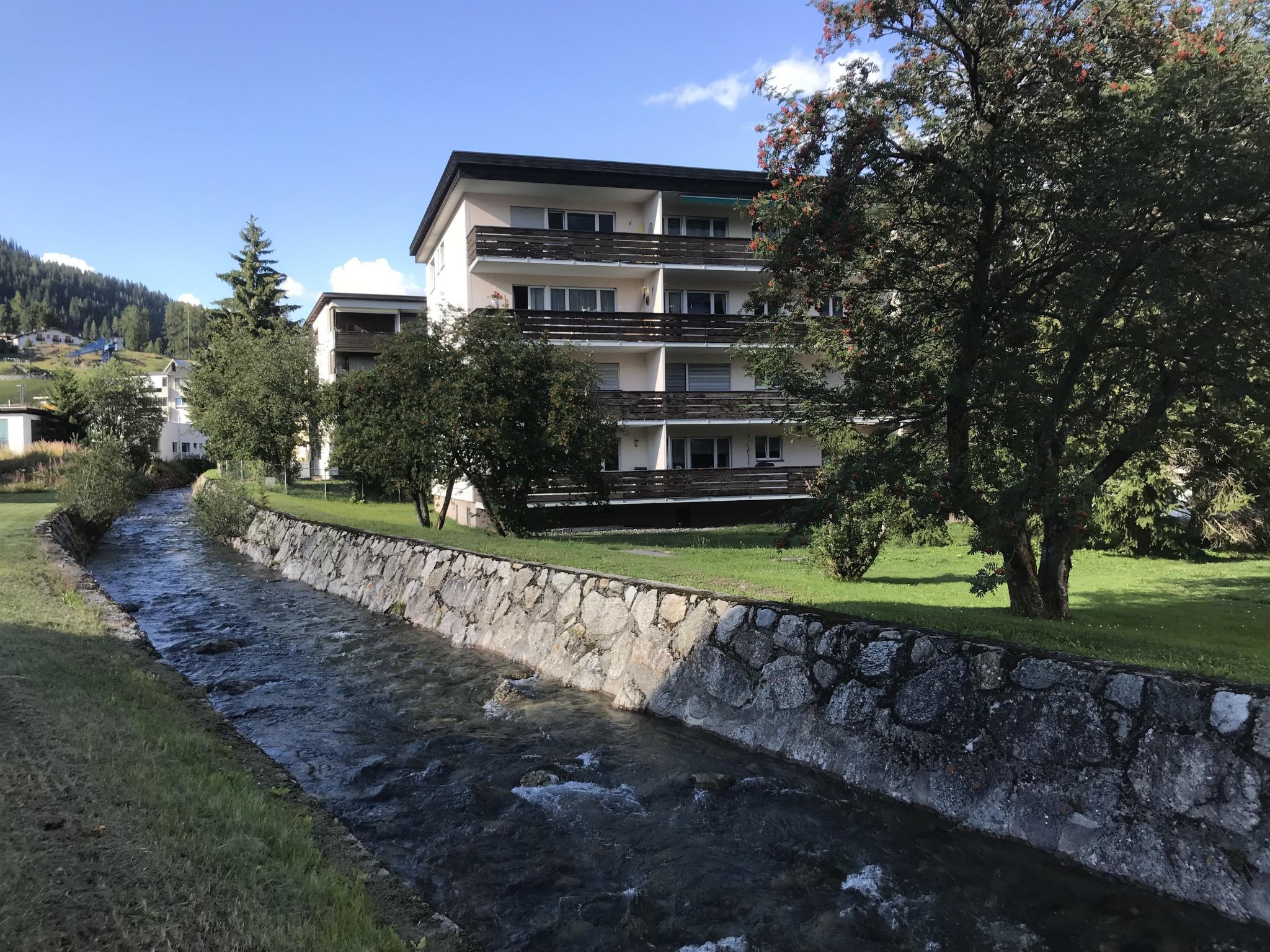 A multi-story building with balconies, a stone retaining wall, and a stream running alongside.