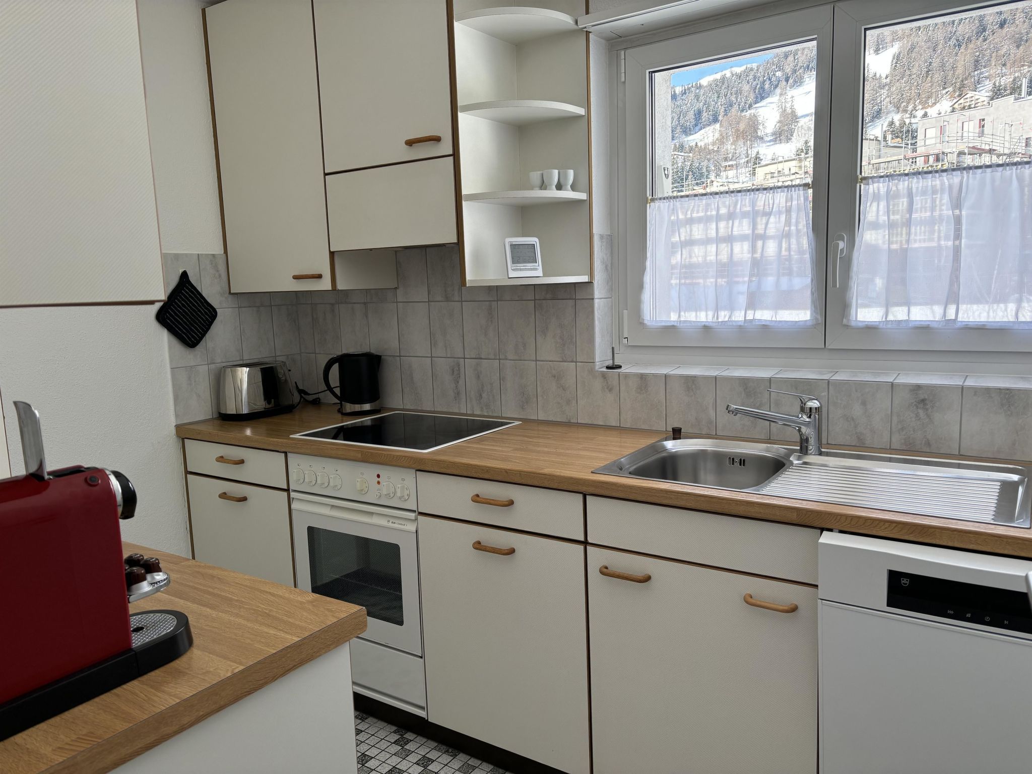 Een kleine keuken met een inbouwoven, een spoelbak en een raam met uitzicht op een besneeerd landschap.