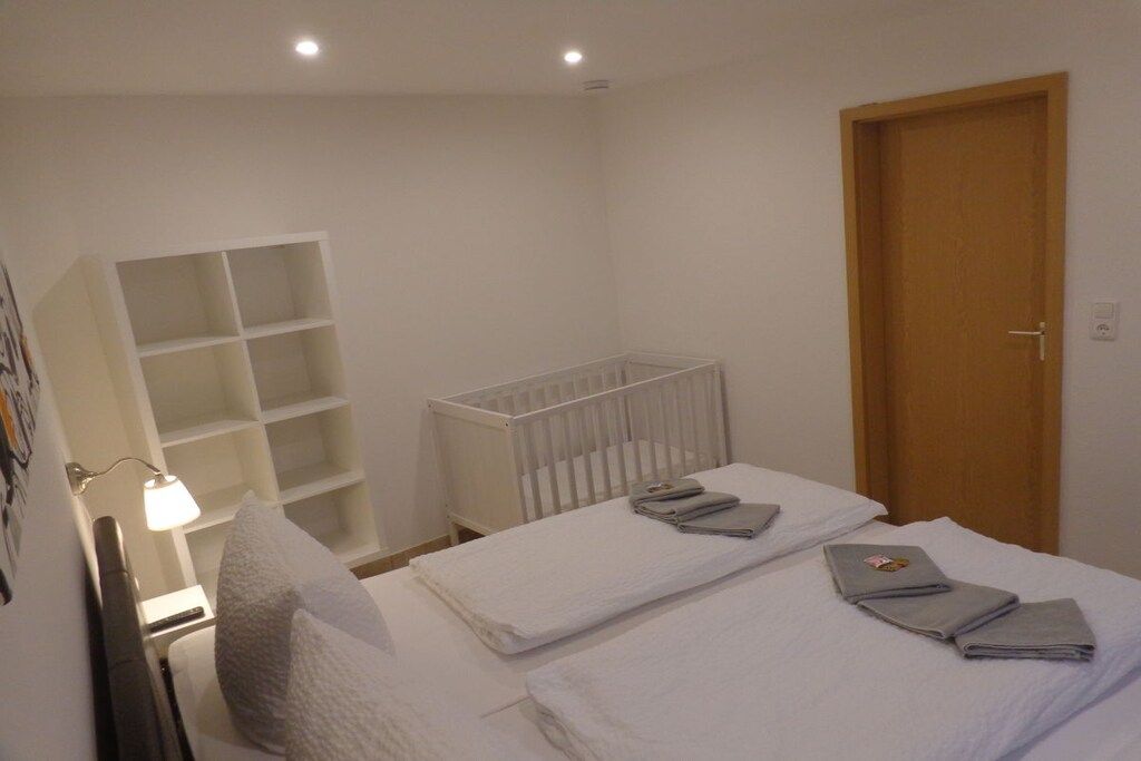 A hotel room with a crib, a bed with neatly folded towels, a bedside lamp, and a wooden door.